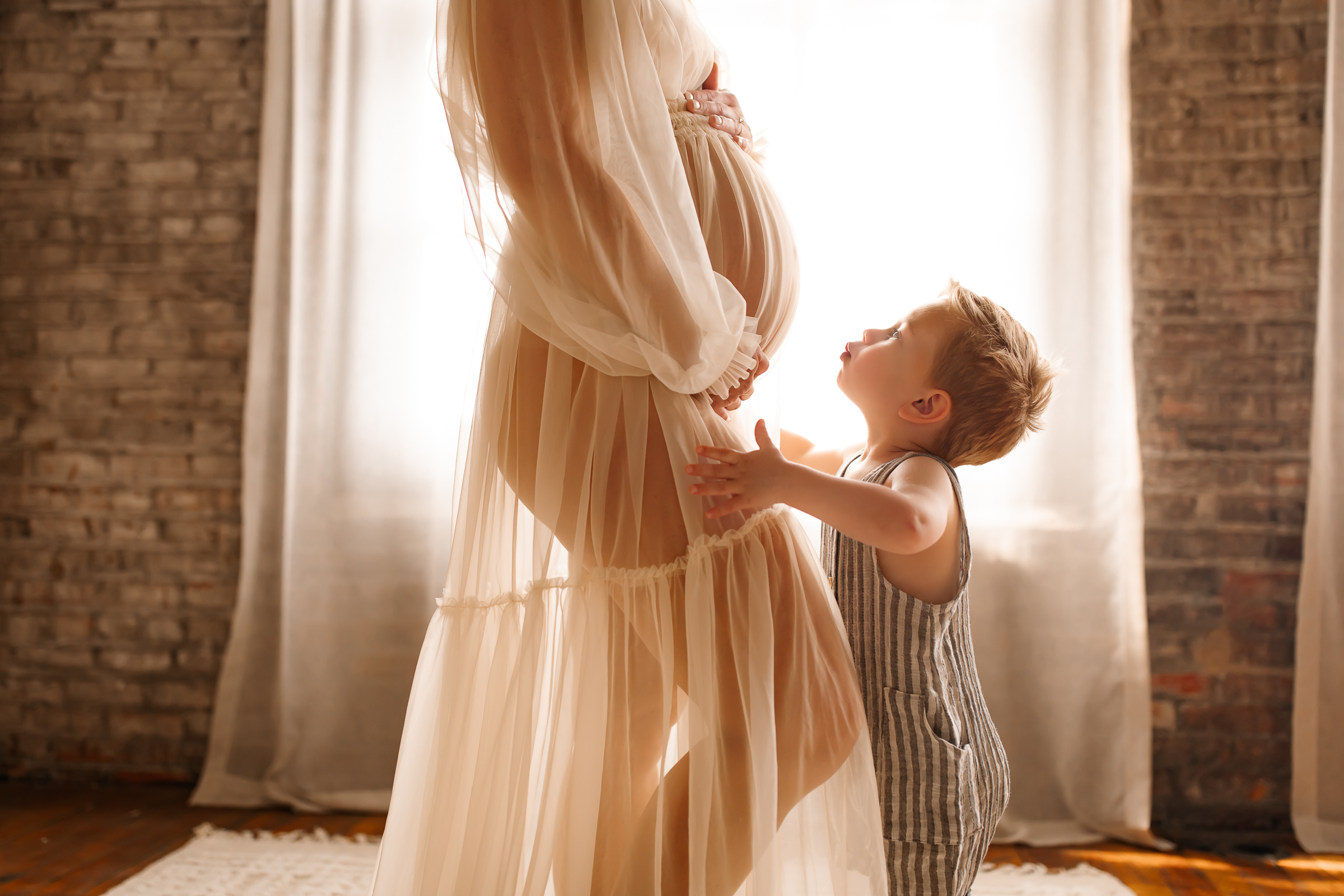Expecting mother posing in front of window with little boy kissing bump
